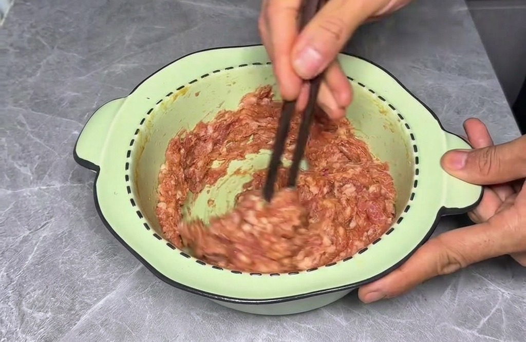 A person using dark chopsticks to vigorously stir seasoned ground meat in a circular motion inside a bowl.
