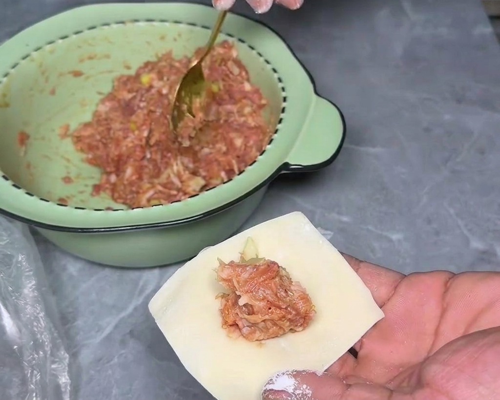 A close-up of a hand holding a single square wonton wrapper with a small mound of meat filling in the center.