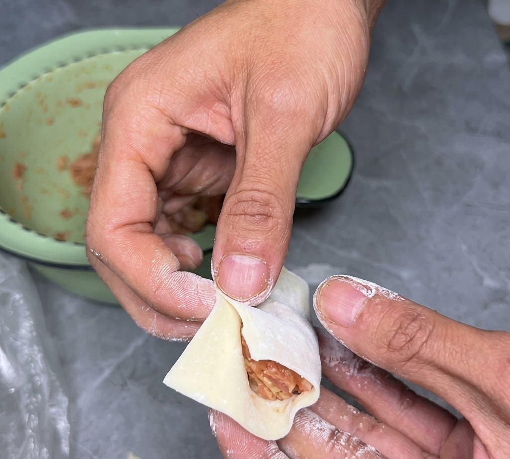 A pair of hands carefully folding a square wonton wrapper in half over a savory meat filling.