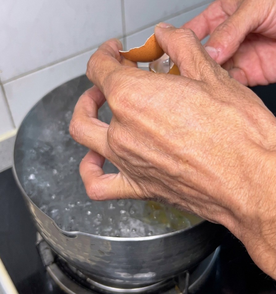 Two hands cracking a brown eggshell over a stainless steel pot of rapidly boiling water.