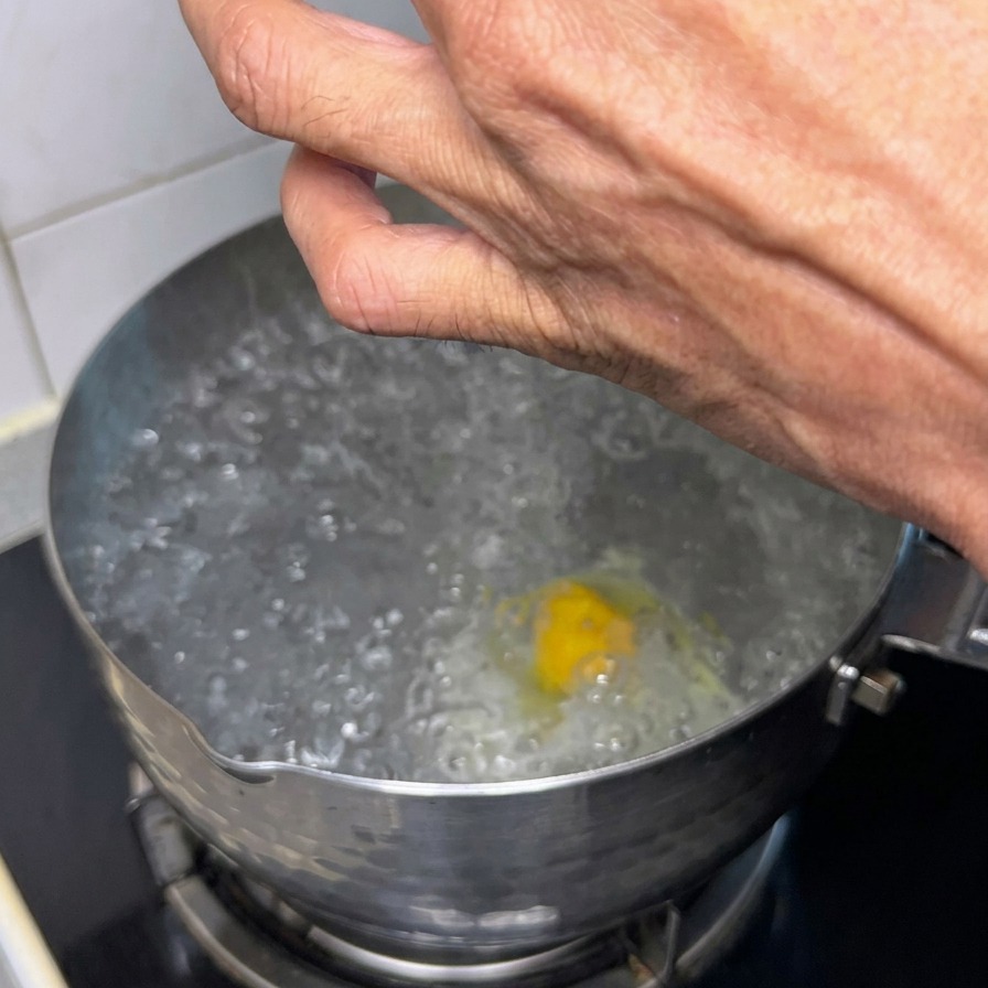 A hand using a spoon to drop a small portion of raw, seasoned meat into a pot of boiling water that already contains a poaching egg.