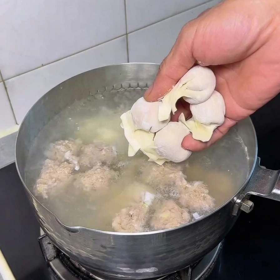 A persons hand adding fresh, raw wontons into a stainless steel pot of boiling water containing meatballs.
