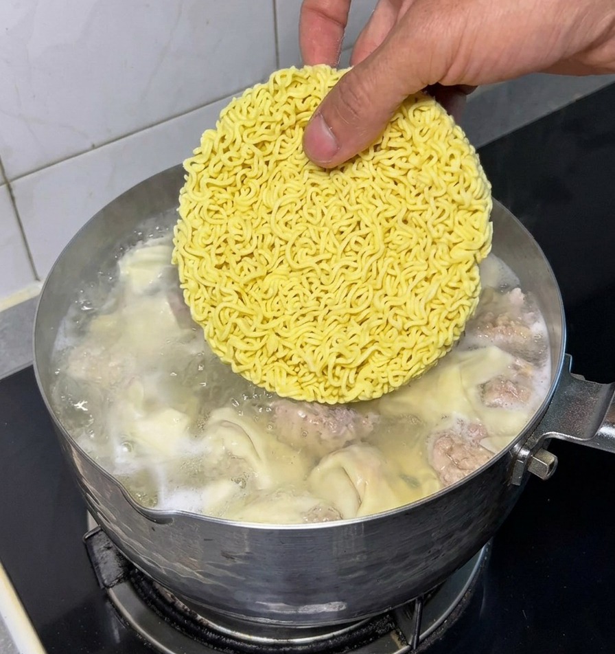 A hand placing a round yellow dried instant noodle block into a pot of boiling water containing cooked wontons and meatballs.