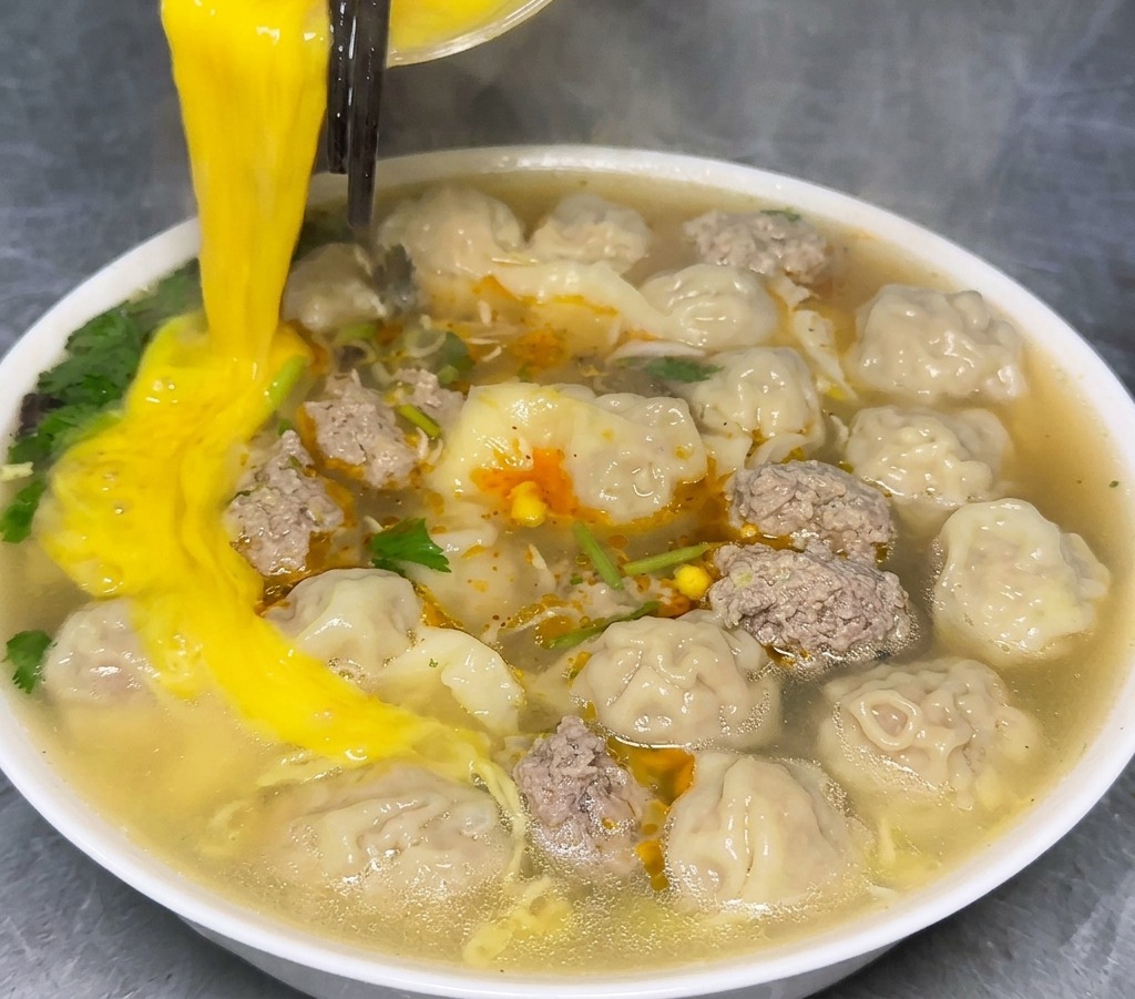 Beaten egg being poured from a glass bowl into a white ceramic bowl filled with steaming wonton noodle soup.