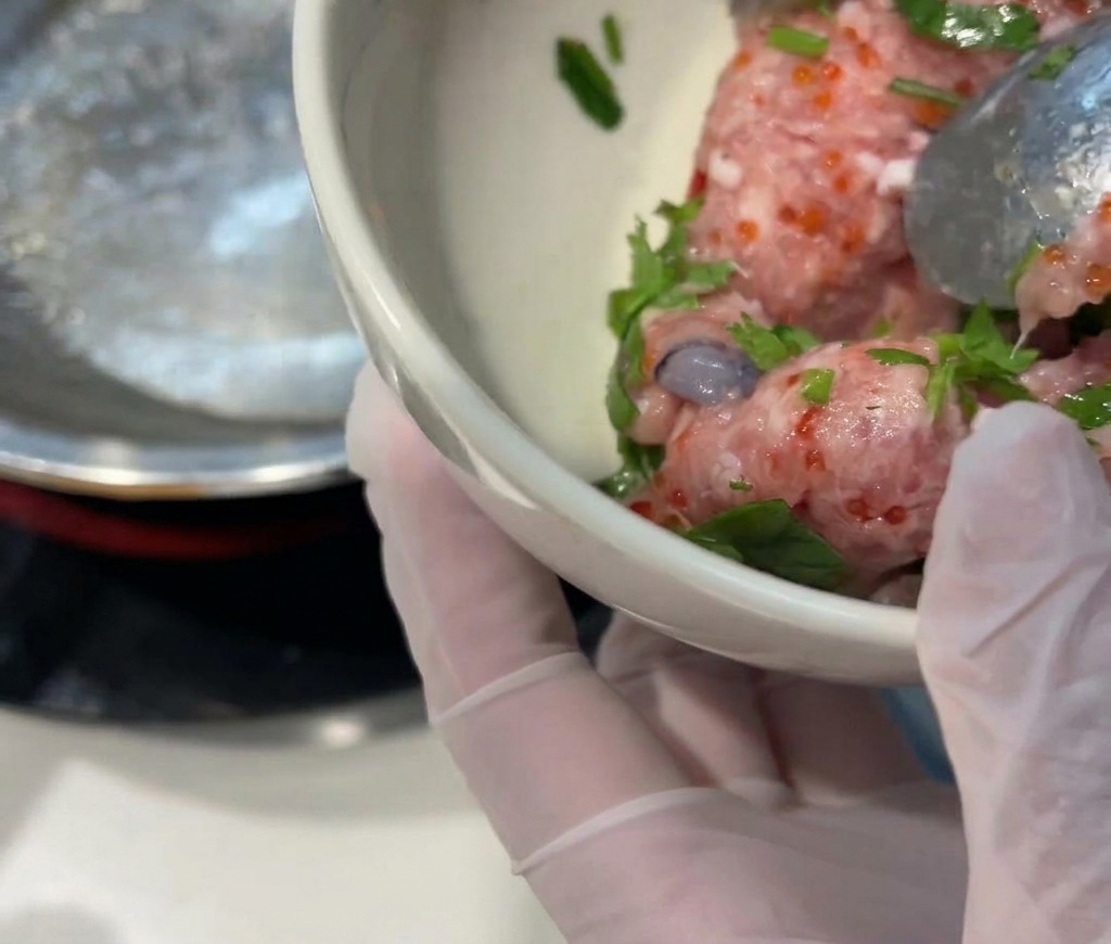 A hand in a clear glove holding a small white bowl filled with raw pink meat filling, bright orange fish roe, green cilantro, and fresh shrimp.