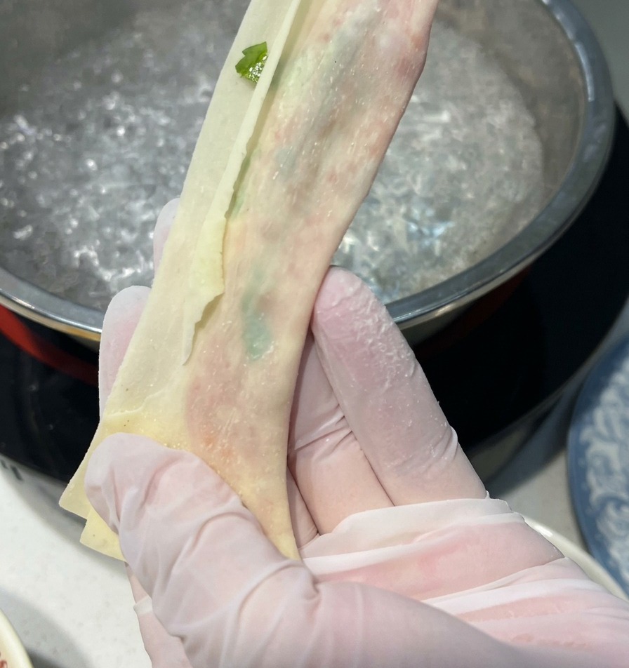Gloved hands stretching a filled wonton wrapper into a long, thin, translucent noodle strip over a pot of boiling water.