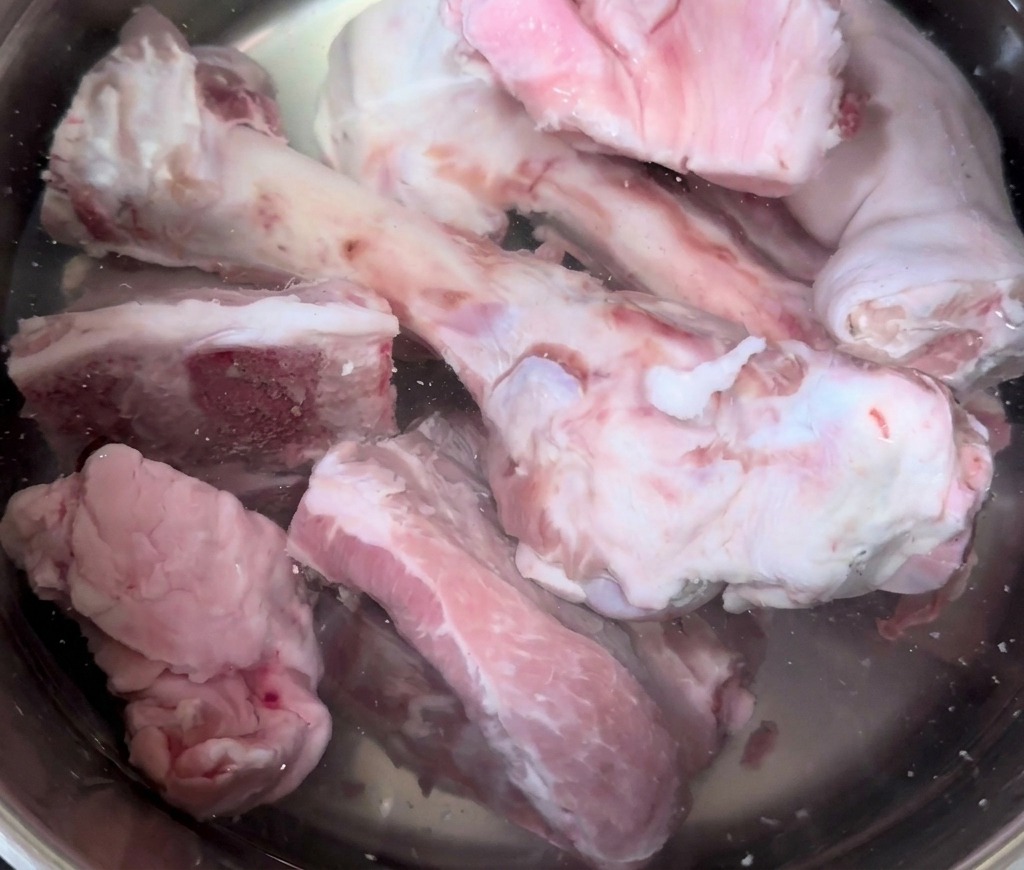 Raw pig bones and trotters being submerged in a large stainless steel pot filled with cold water.