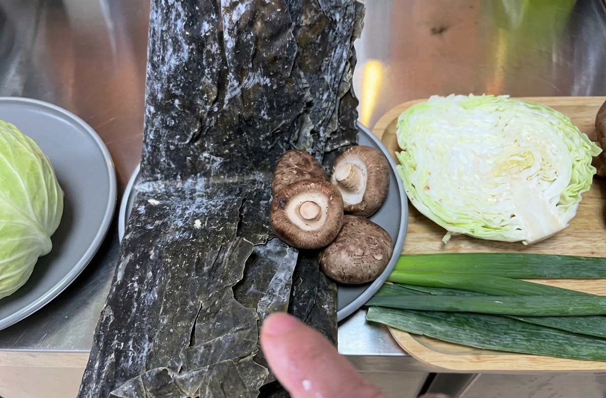 A variety of fresh ingredients including cabbage, kombu seaweed, and shiitake mushrooms arranged on a kitchen counter for preparation.