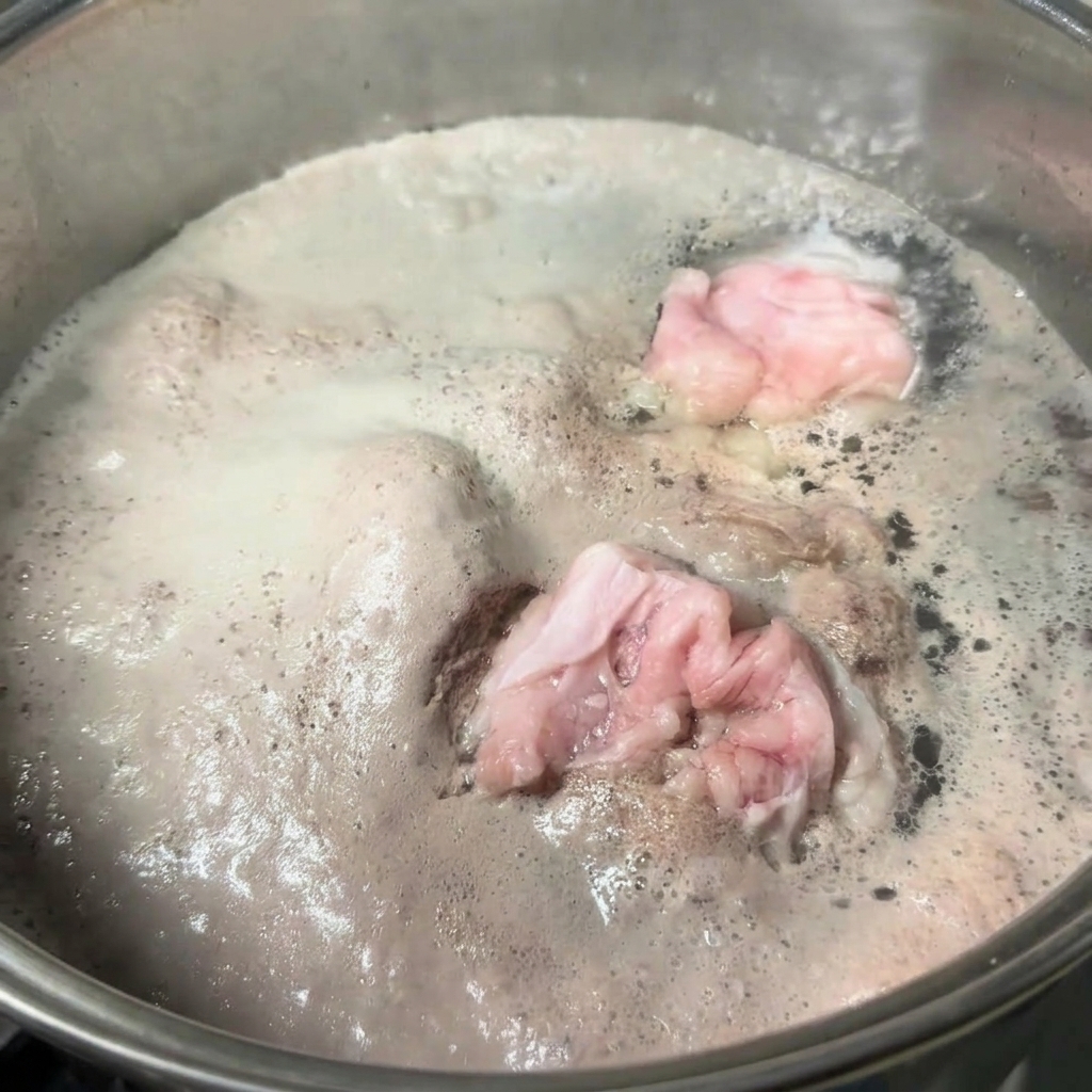 A stainless steel pot filled with boiling water and pork bones, with a thick layer of grey foam floating on the surface.