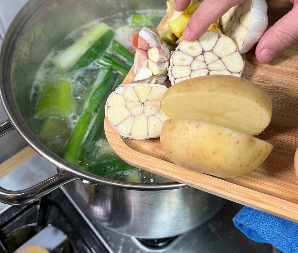 A hand adding halved garlic bulbs and potatoes from a wooden board into a steaming pot of bone broth.