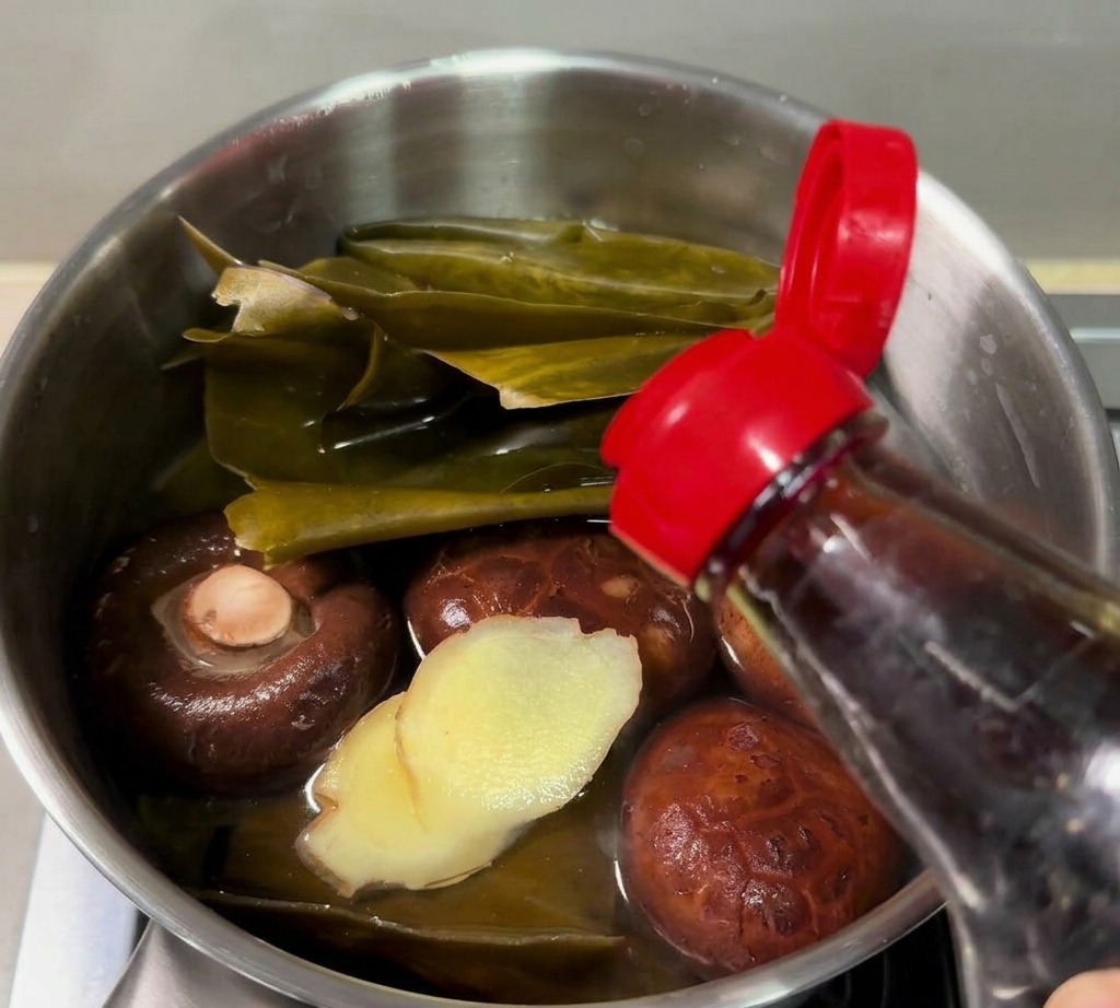 Fish sauce being poured from a bottle into a small saucepan containing kombu, shiitake mushrooms, and sliced ginger.