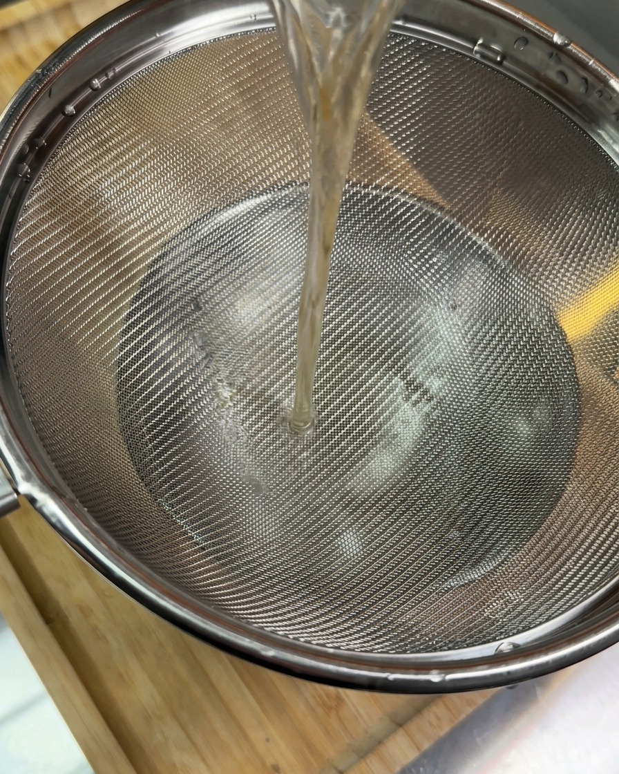 A stream of dark brown liquid being poured through a metal mesh strainer into a bowl below.