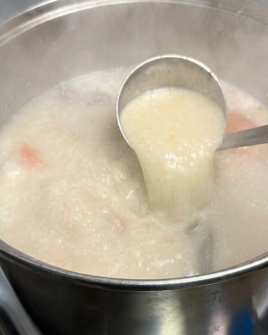 A metal ladle lifting a scoop of thick opaque milky white tonkotsu broth from a large simmering stockpot.