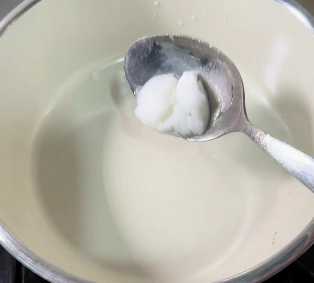A metal spoon holding a dollop of white, solid pork lard over the center of a small cream-colored saucepan.