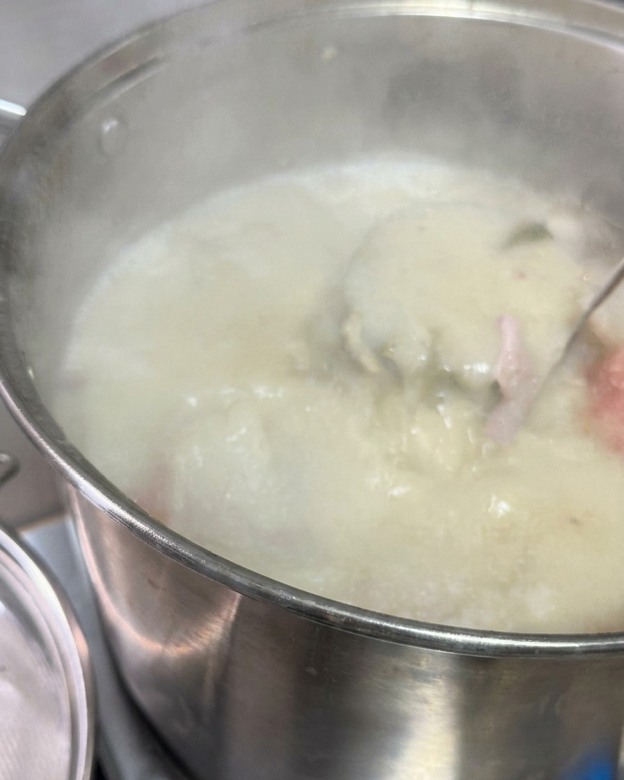 Milky white pork broth being poured through a metal sieve into a pot containing a dark sauce and lard.