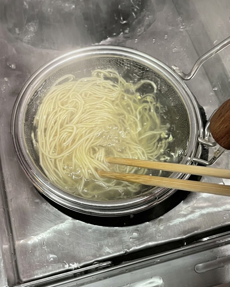 A bundle of thin, pale wheat noodles being lifted out of boiling water inside a circular mesh noodle strainer using wooden chopsticks.