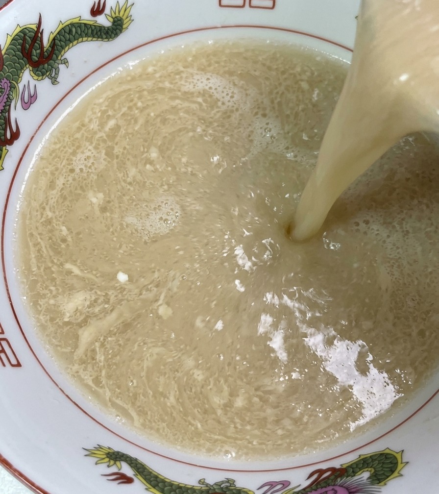 Steaming, ivory-colored pork bone broth being poured from a ladle into a decorated ramen bowl, showing the rich and silky texture of the liquid.