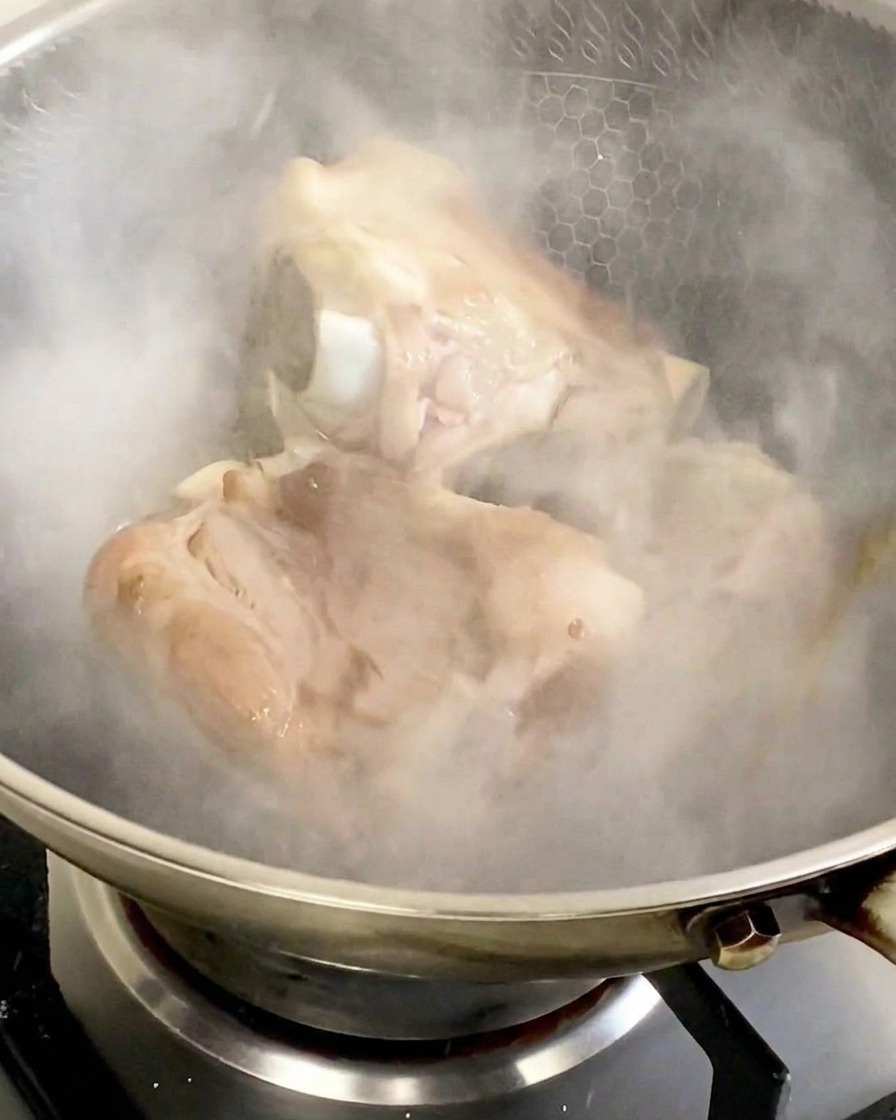 Pork bones in a stainless steel wok being enveloped in a thick cloud of white steam after the addition of cooking wine.