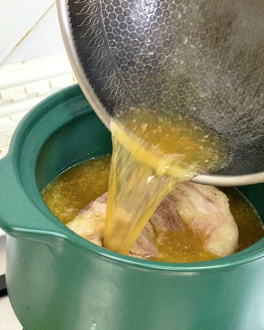 Liquid and oil from a frying pan being poured into a green clay pot containing seared pork bones.