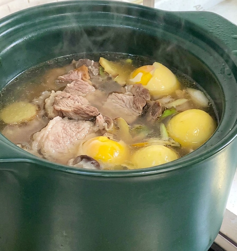 A green clay pot containing a bubbling pork bone soup with visible chunks of corn and yam simmering on the stove.