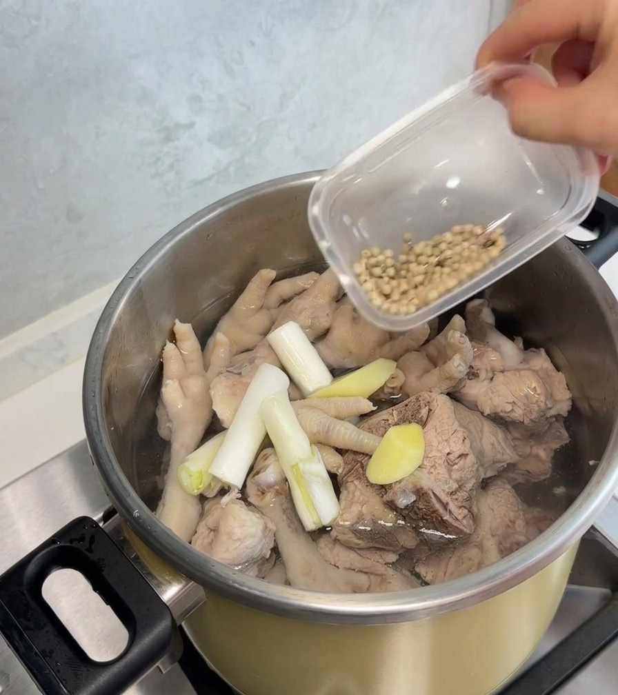 A hand pouring white peppercorns from a small plastic container into a stainless steel pot filled with blanched chicken feet, pork bones, scallion stalks, and ginger slices.
