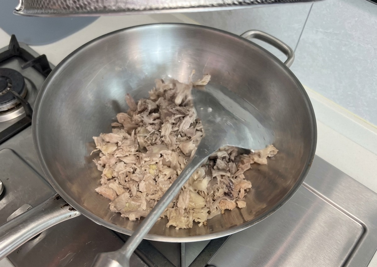 Shredded cooked meat scraps being stir-fried and mashed with a metal spatula in a large stainless steel wok on a stovetop.