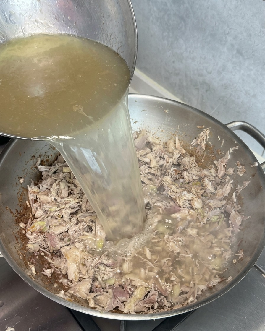 A ladle pouring clear golden broth into a wok filled with browned meat scraps, creating a cloudy white reaction upon contact.