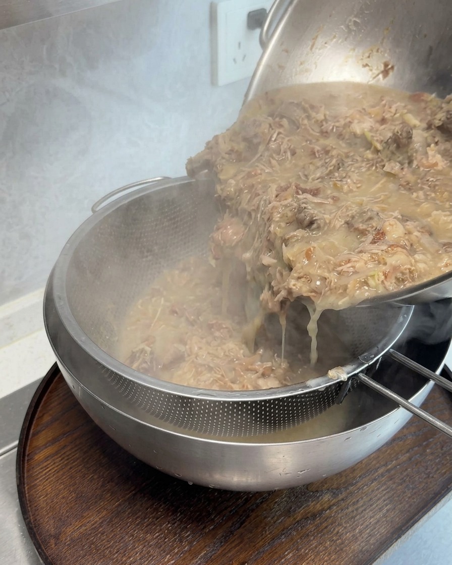 Thick, concentrated bone broth being poured through a fine mesh strainer to remove meat scraps.