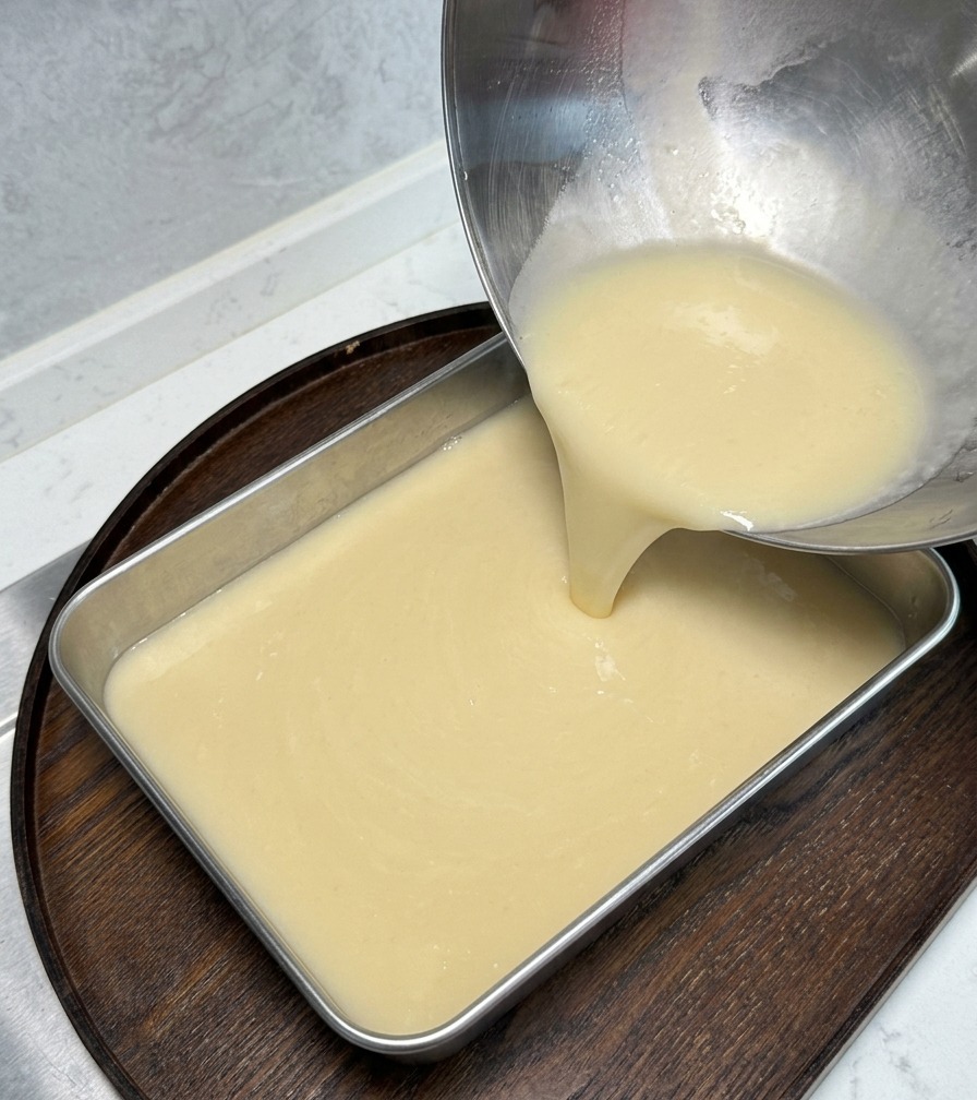 A thick, cream-colored broth being poured from a stainless steel bowl into a rectangular metal tray placed on a wooden board.