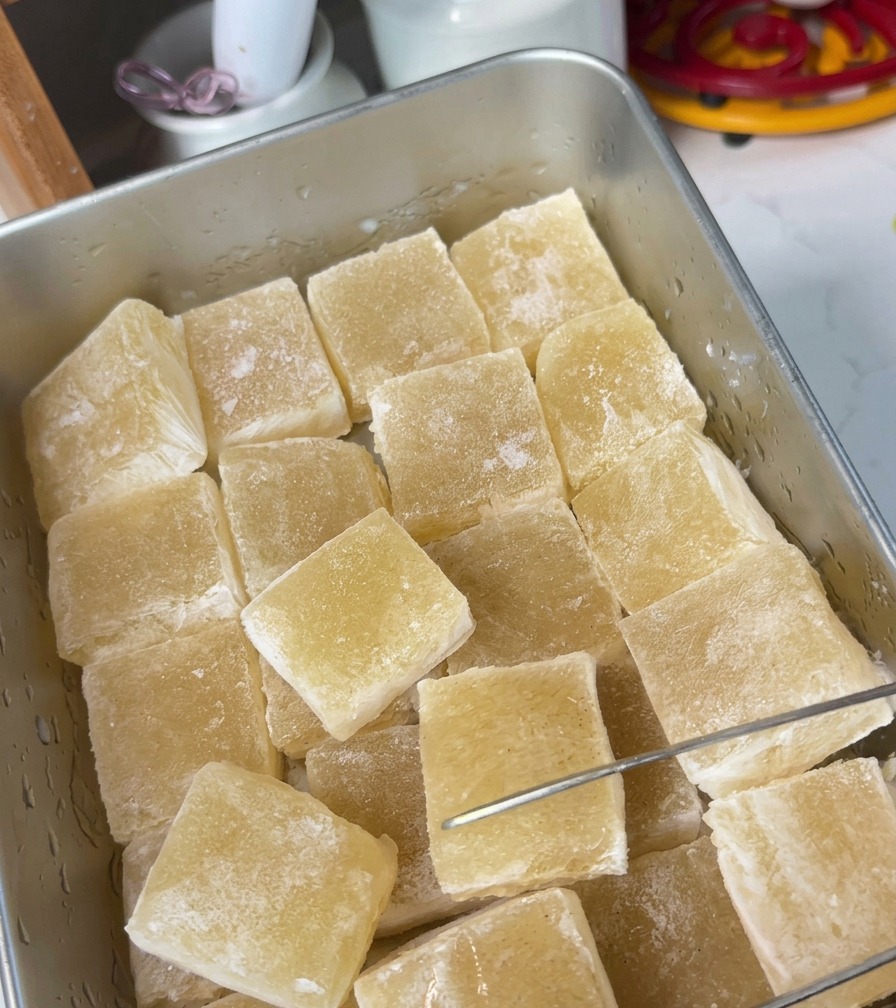 A metal tray filled with square, translucent yellow broth jelly cubes being sliced by a metal tool.