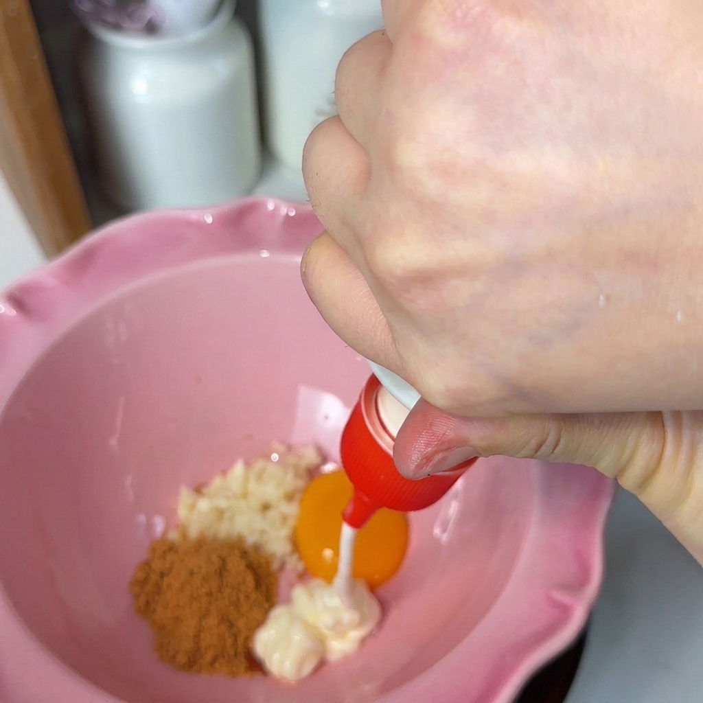 White mayonnaise being squeezed from a red nozzle into a pink fluted bowl containing minced garlic, a bright orange egg yolk, and brown seasoning powder.