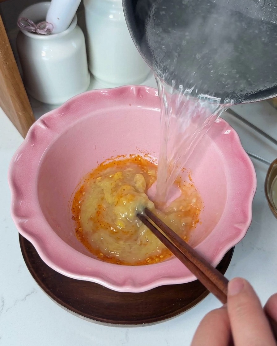 Hot clear liquid being poured from a pot into a pink bowl containing a creamy yellow seasoning paste, stirred with wooden chopsticks.