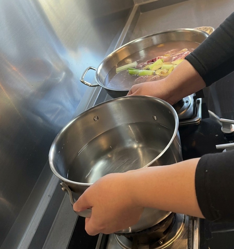 A pair of hands placing a stainless steel pot filled with clean water onto a gas stove burner.