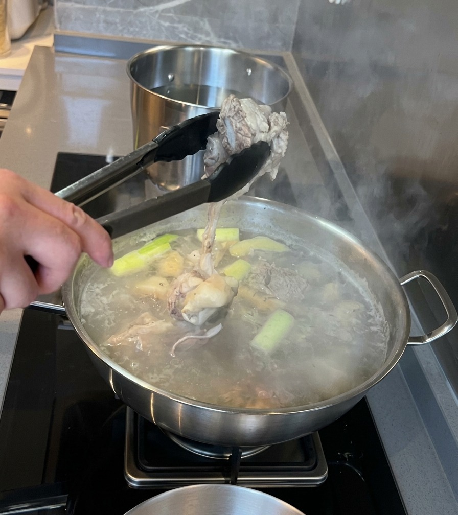 Metal tongs lifting a piece of blanched pork marrow bone out of a pot of simmering water with aromatics.