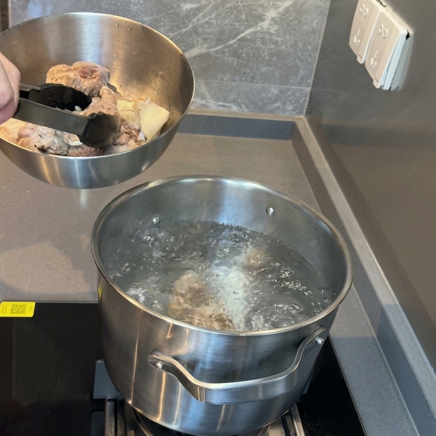 A metal bowl filled with blanched pork bones being held over a pot of boiling water, ready for transfer.