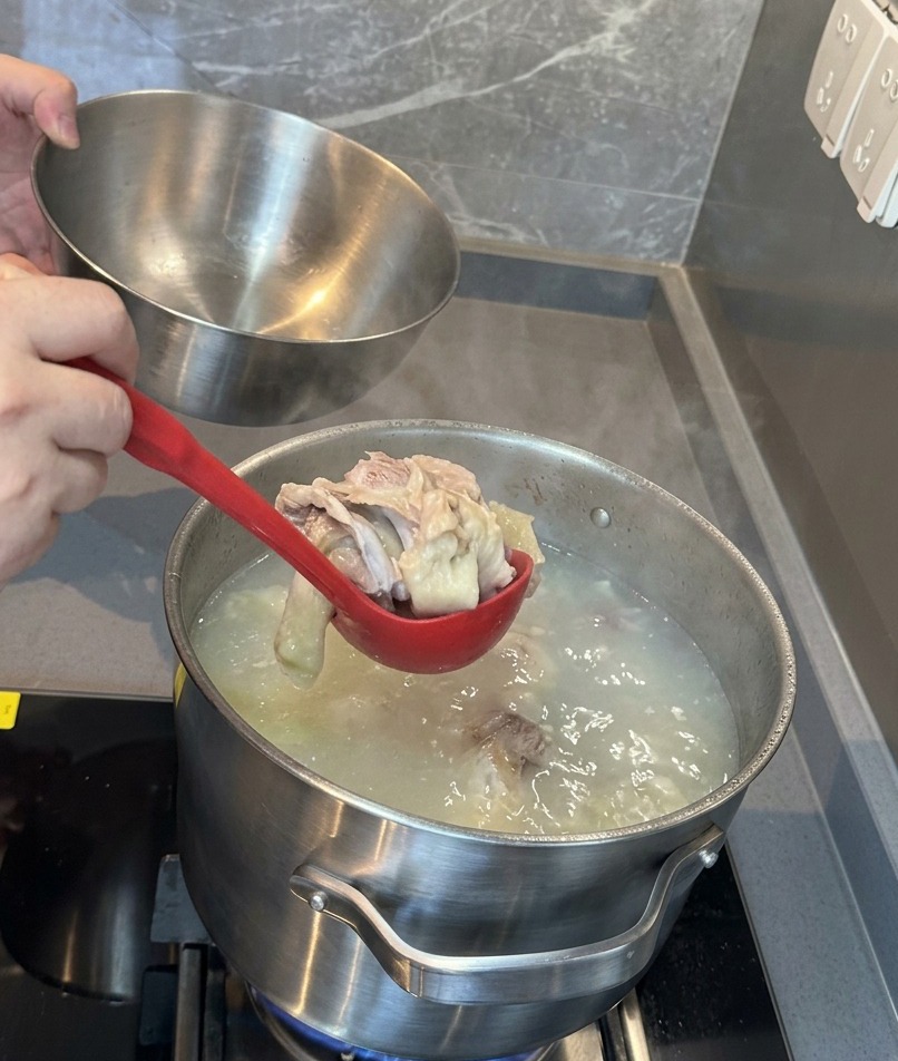 A red ladle lifting softened pork bones and meat out of a milky white broth into a stainless steel bowl.