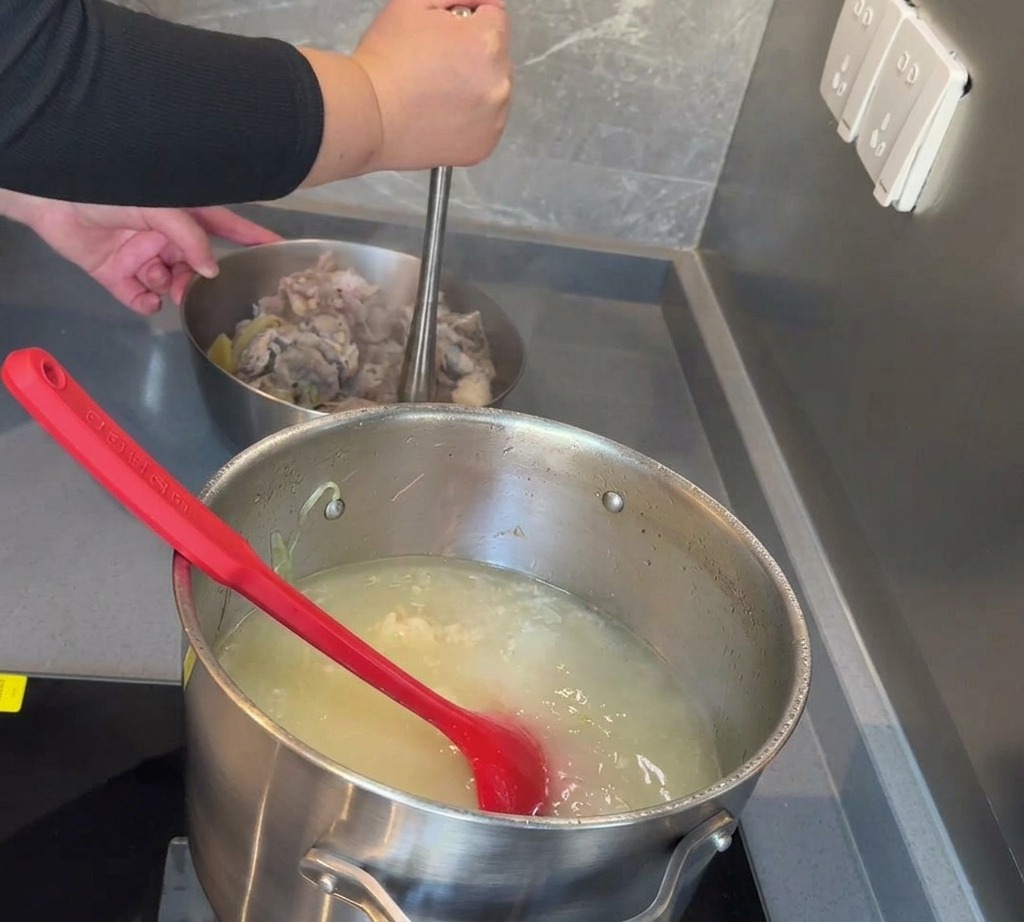 A hand using a metal masher to crush pieces of boiled pork and marrow in a stainless steel bowl on a counter.