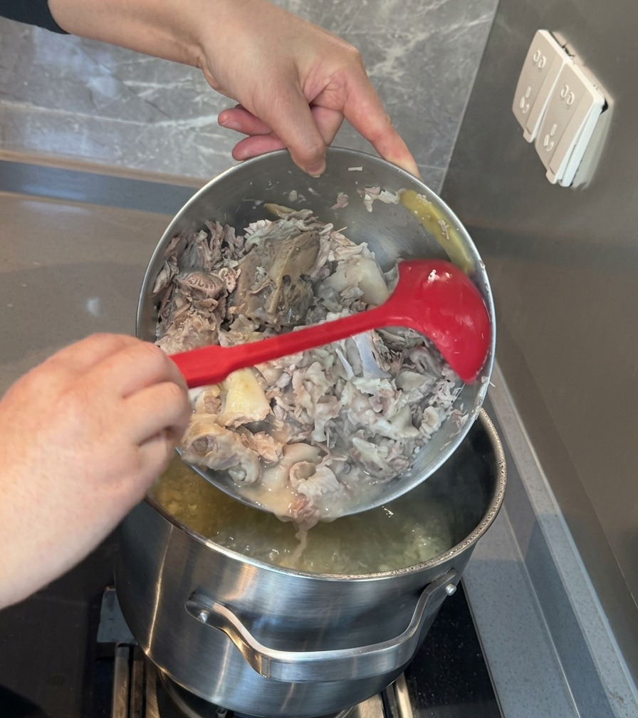 The contents of a metal bowl, consisting of mashed pork meat and bones, being poured back into a large stockpot of white broth.