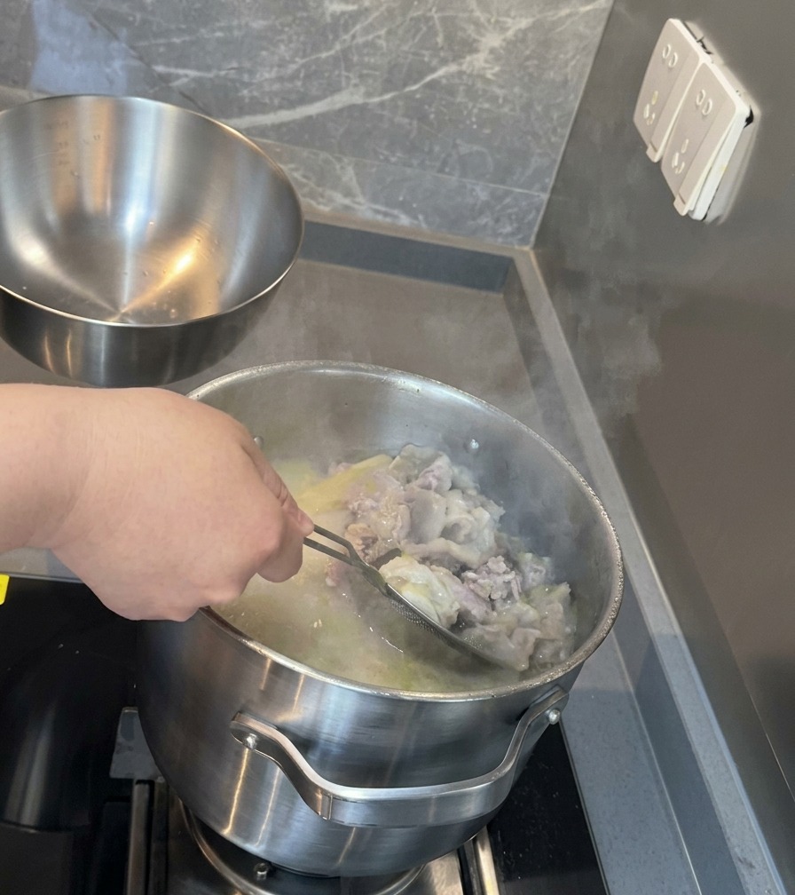 A stainless steel mesh strainer filtering out bits of bone and meat from a deep pot of milky white pork soup.