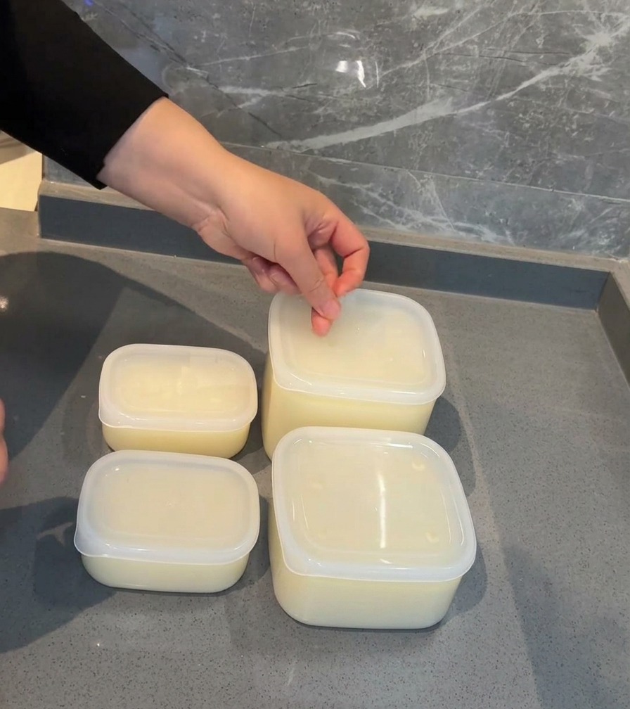 A hand applying a white date label to the lid of a clear plastic container filled with pork broth.