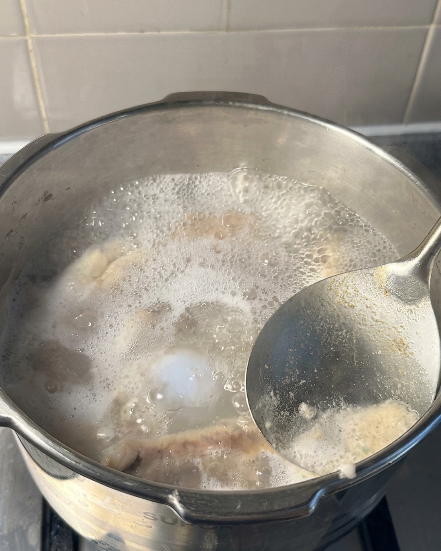 A close-up of a metal ladle lifting white and grey foam from the surface of boiling water inside a pressure cooker.