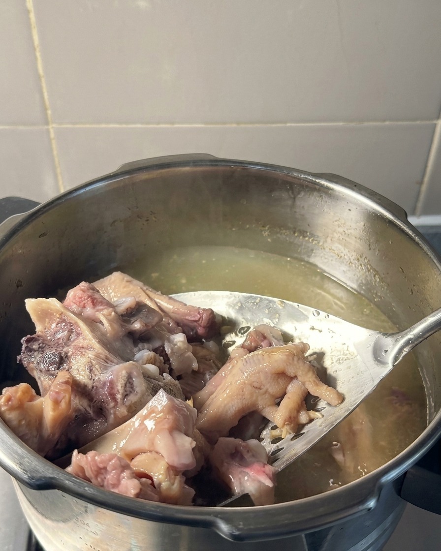A slotted metal spatula lifting tender, cooked pork and chicken feet out of a pot of pale yellow broth.