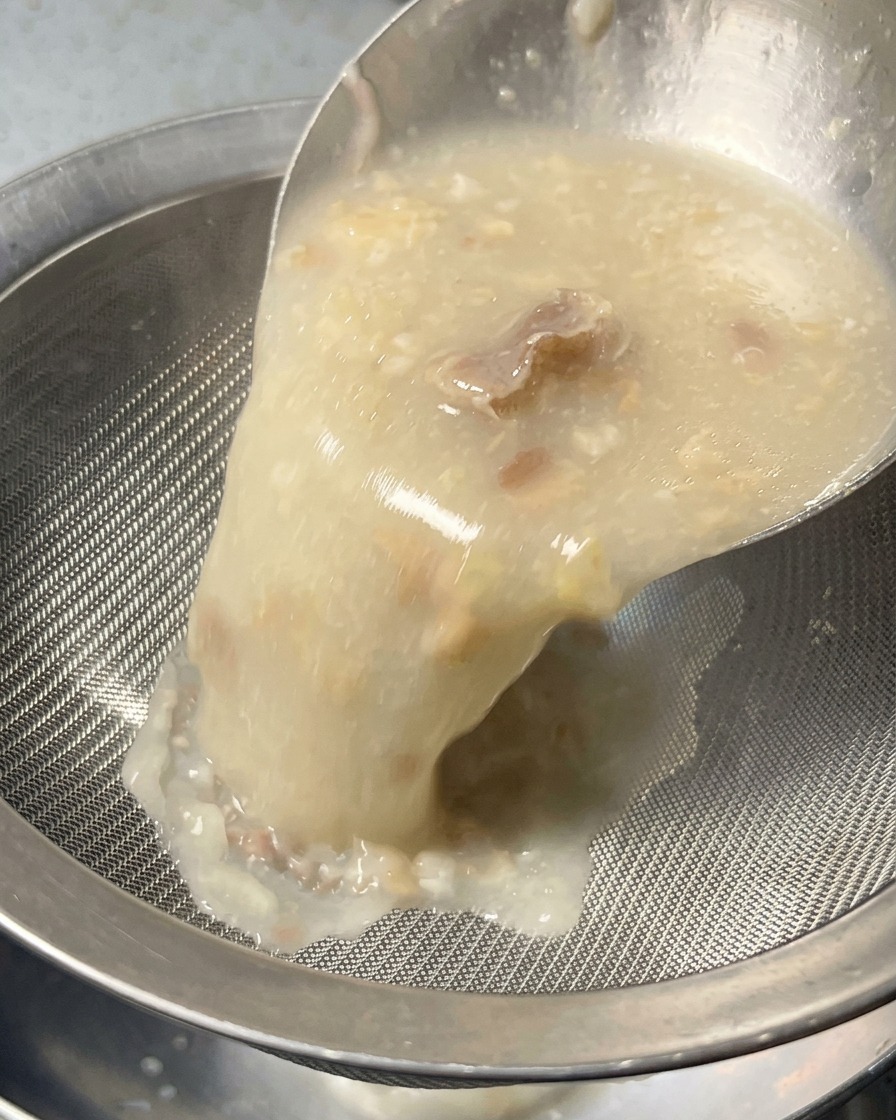 A thick, opaque white broth being poured through a silver mesh strainer into a bowl, filtering out solid bits.