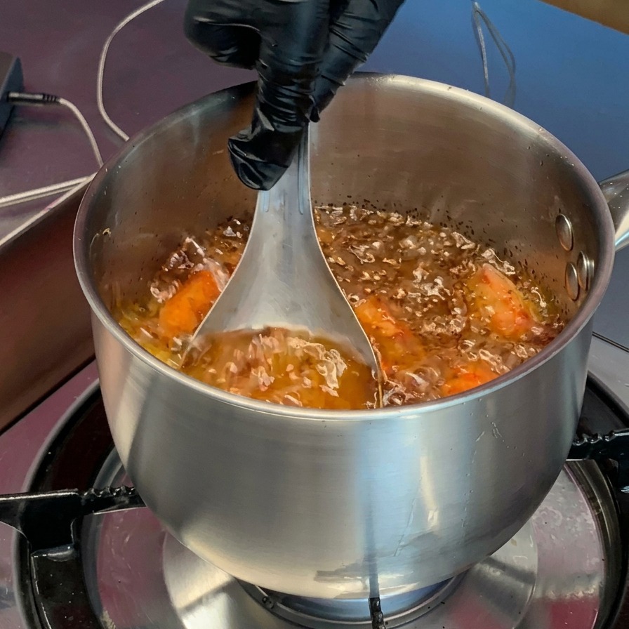 A gloved hand pressing orange, cooked shrimp heads with a metal ladle into the bubbling hot oil inside a pot.