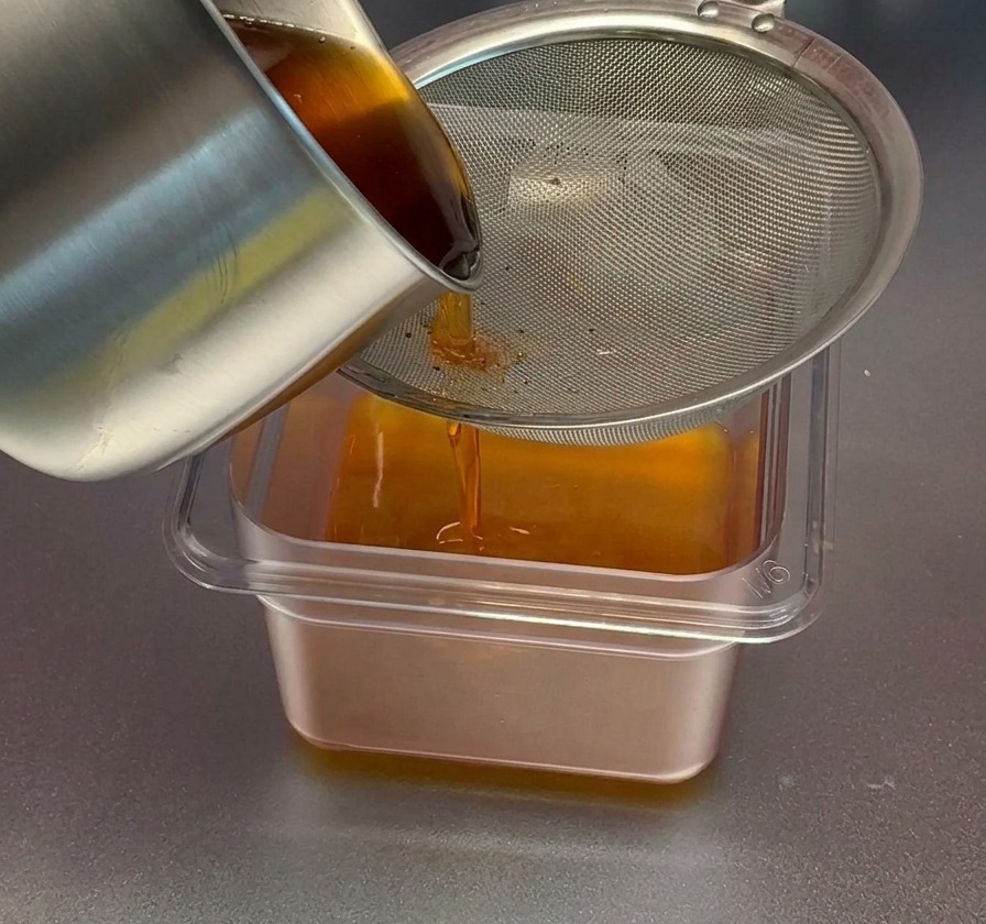 Pouring reddish-orange shrimp oil from a stainless steel pot through a fine metal mesh strainer into a clear rectangular container.