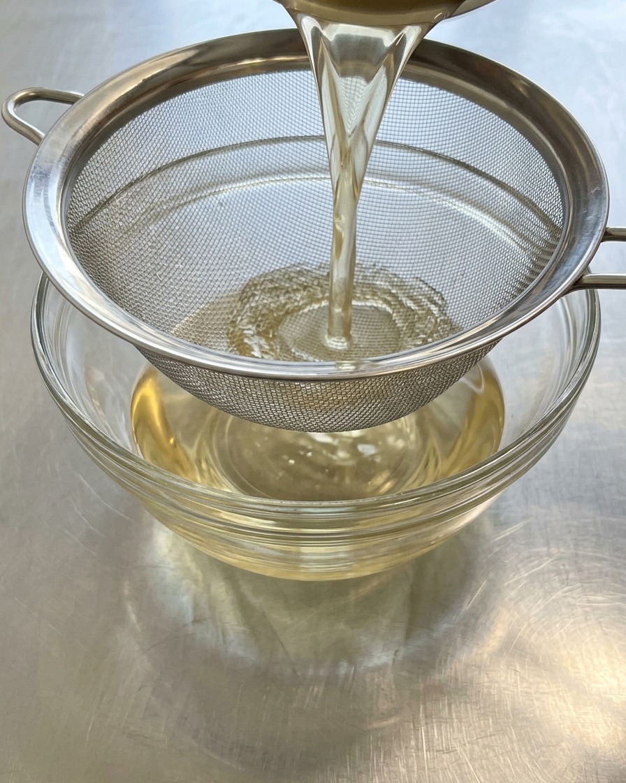 Golden-brown bonito broth being poured through a metal strainer into a clear glass bowlcite: 60.