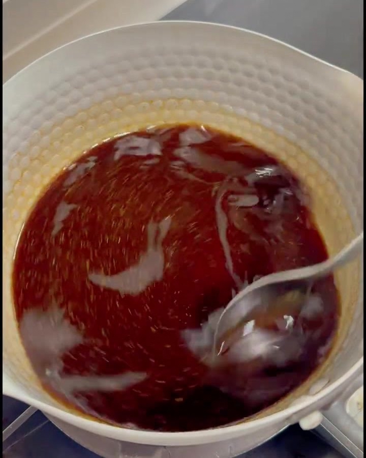 A dark shimmering soy-based seasoning liquid being stirred with a metal spoon in a white hammered-metal pot on a stove.