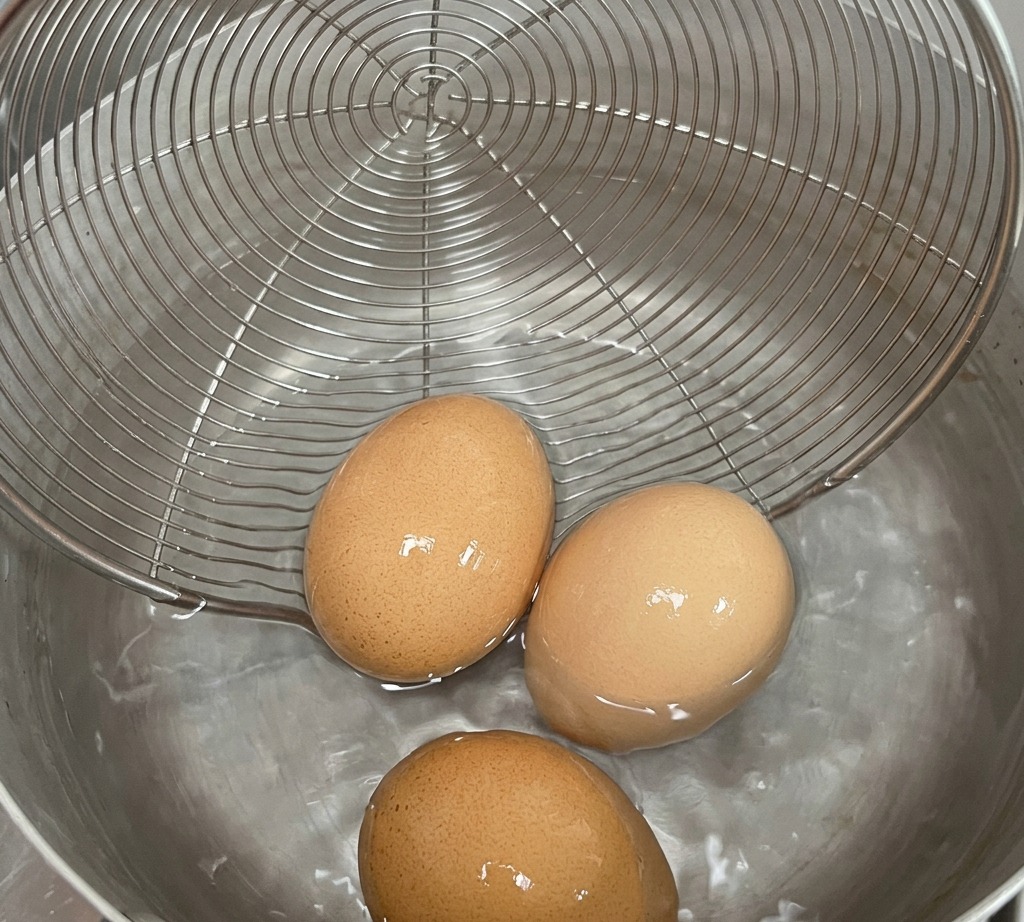 Three brown eggs in a wire mesh strainer being lowered into a pot of hot water.