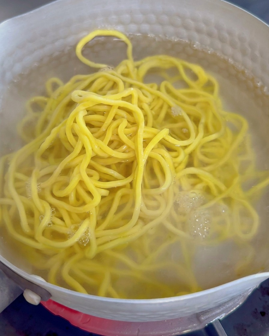Yellow thick ramen noodles boiling in a pot of water with white foam and steam visible.