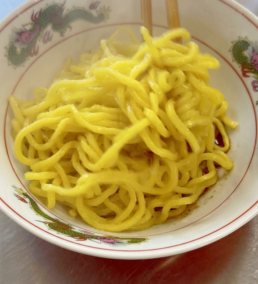 A close-up shot of thick, curly yellow ramen noodles steaming in a white bowl, with a dark soy sauce base visible at the bottom.